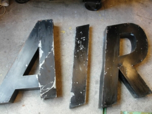 De grandes lettres noires vieillies épelant « AIR » reposaient sur une surface de béton rugueuse, évoquant un attrait vintage rappelant les marchés de brocante.