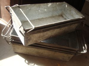 Trois conteneurs rectangulaires métalliques empilés avec poignées, dégageant un charme rétro, placés sur un sol en béton au soleil.
