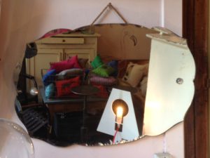 Un miroir décoratif vintage reflétant une pièce confortable avec des coussins colorés, des meubles rétro et une lampe lumineuse.