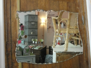Miroir déformé sur une porte en bois vintage reflétant une chaise en osier, des armoires métalliques et divers objets Brocante dans une pièce.