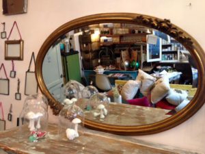 Miroir ovale vintage avec cadre doré orné reflétant une pièce avec des étagères, des chaises et des coussins ; petits objets de décoration sur la table.