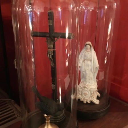 Deux cloches de présentation en verre de la collection Globes de Mariage 1930 : l'une présentant un crucifix et un corbeau, l'autre mettant en valeur une statue de la Vierge Marie, le tout posé sur une surface en bois évoquant la brocante 93. Brocante 93