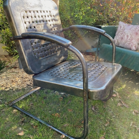 Le Fauteuil de Jardin Américain 1950 VENDU, une chaise rétro vintage en métal avec accoudoirs et dossier en résille, trône sur l'herbe d'un jardin. A proximité, des plantes et un banc vert créent une charmante ambiance brocante 93. Brocante 93