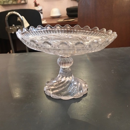 La Coupe en Verre 1950 Motifs Arabesques, dotée d'un bord ondulé et d'une tige torsadée qui respire le charme brocante, est élégamment placée sur une table sombre au milieu d'un éventail d'objets d'arrière-plan dans la pièce. Brocante 93