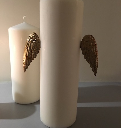 Deux bougies piliers blanches Ailes Murales, ornées d'ornements d'ailes dorées sur leurs côtés, reposent sur une surface grise, évoquant une charmante esthétique brocante 93. Brocante 93