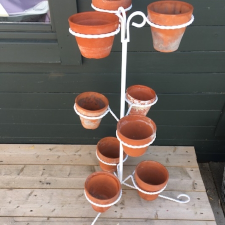 Un présentoir en métal blanc, connu sous le nom de Porte-Pots Fer Forgé 1950, supportant plusieurs pots en terre cuite vides sur une terrasse en bois avec un mur vert foncé en arrière-plan, rappelant les charmantes vitrines vintage de la brocante 93. Brocante 93