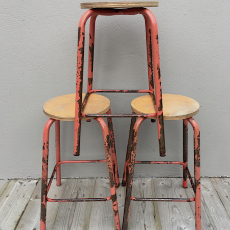 Trois tabourets vintage en bois rouge des années 1960, étiquetés «Tabourets d'Usine 1960 VENDU», sont empilés les uns contre les autres contre un mur gris sur un plancher en bois.

 Brocante 93