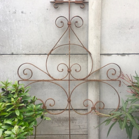 Un treillis métallique rouillé et orné de motifs tourbillonnants se dresse contre un mur de béton, entouré de plantes vertes luxuriantes, évoquant le charme de la Croix de Calvaire 1940. Brocante 93