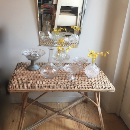 Une table en osier et rotin des années 60 ornée de verrerie et de fleurs jaunes, un miroir vintage au mur reflétant une pièce au mobilier rétro. Brocante 93