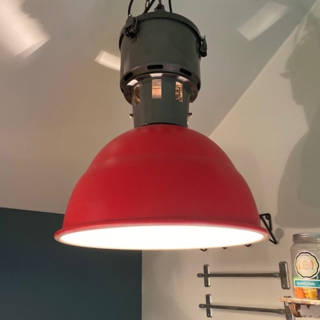 La Suspension Industrielle 1960 VENDU, luminaire suspendu de style industriel de couleur rouge au design rétro, est suspendue au plafond dans une pièce aux murs et étagères sombres, créant une ambiance vintage. Brocante 93