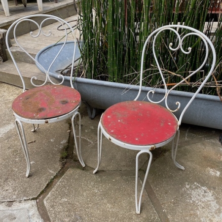 Deux Chaises Fer Forgé 1950 aux assises rouges patinées sont posées sur un patio en pierre près d'une zone de plantation avec de la verdure haute et fine, ajoutant une touche de charme rétro à la scène. Brocante 93