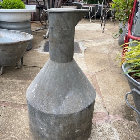 Le Broc en Galva 1960, avec son look vintage et patiné, se dresse debout sur une terrasse extérieure entourée de plantes en pot et d'outils de jardin, ajoutant une charmante touche rétro à la scène. Brocante 93