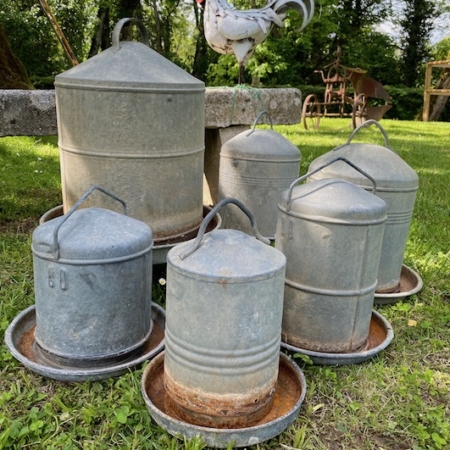 Six mangeoires à poulets vintage en métal "Distributeurs de Grain 1960" de différentes tailles reposent sur l'herbe, avec une statue de coq décorative en arrière-plan, donnant une charmante ambiance brocante 93. Brocante 93