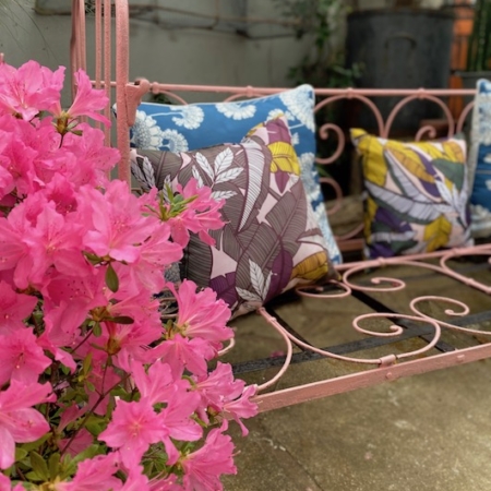 Un lit Enfant Fer Forgé vintage des années 1950 avec des coussins floraux colorés, évoquant un charme rétro, se trouve à côté de fleurs roses vibrantes dans un décor de jardin extérieur. Brocante 93