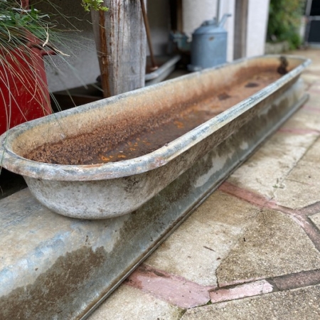 Une Auge à Bétail 1960 GM, remplie d'eau trouble, repose sur un sol pavé de pierres à proximité d'outils et de plantes de jardin d'époque. Brocante 93