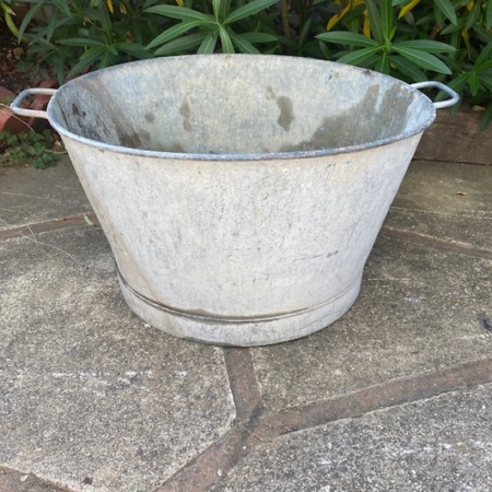 La Bassine Ronde 1960 PM, un grand seau en métal patiné avec poignées, ajoute une touche vintage parfaite au patio en pierre, entouré de plantes à feuilles vertes en arrière-plan. Brocante 93