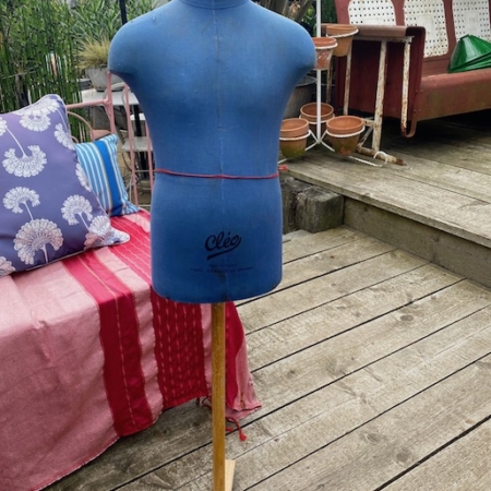 Le Mannequin de Couture Enfant 1960, un mannequin de couture bleu vintage sur un support en bois, est placé sur une terrasse avec un coin salon extérieur et des plantes en pot en arrière-plan. Brocante 93