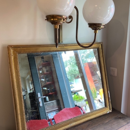 Un Miroir Perlé XXème VENDU repose contre un mur sous une applique à double globe ; une fenêtre et des chaises rouges s'y reflètent, ajoutant une touche rétro à l'espace. Brocante 93