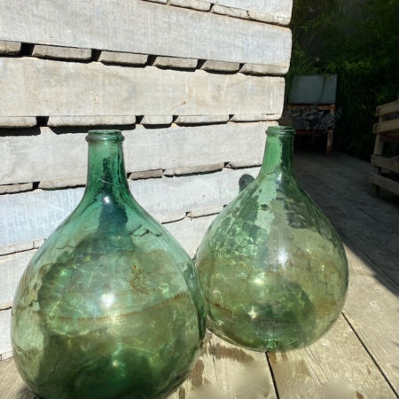 Deux bouteilles en verre vert vintage Dame Jeanne 1960 PM VENDU, de forme bulbeuse, reposent sur un plancher en bois contre un mur de briques au soleil. Brocante 93