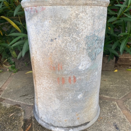 Le Fût Industriel 1960, une jardinière cylindrique en pierre patinée par les intempéries, se dresse sur une allée carrelée, dégageant un charme vintage avec des plantes vertes feuillues en arrière-plan. Brocante 93