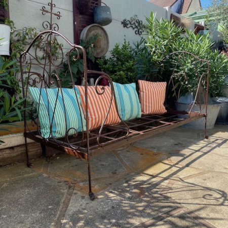 Un banc vintage Lit Fer Forgé 1940 VENDU orné de coussins rayés colorés dans un jardin patio ensoleillé, entouré de plantes en pot. Brocante 93