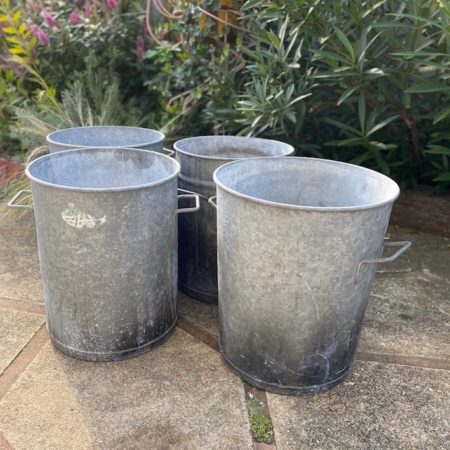 Quatre poubelles métalliques Lessiveuses 1960 à poignées sont disposées sur un patio en pierre, entourées de plantes vertes luxuriantes en arrière-plan. Brocante 93