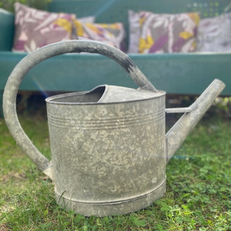 Un arrosoir Arrosoir Galva 1960 repose sur l'herbe avec un banc vert et des coussins colorés en arrière-plan flou, évoquant un charme vintage rappelant un décor de brocante. Brocante 93