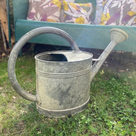 Un arrosoir en métal patiné Arrosoir Galva 1960 VENDU sur l'herbe avec un banc bleu sarcelle et des coussins colorés en arrière-plan, dégageant une charmante sensation vintage. Brocante 93