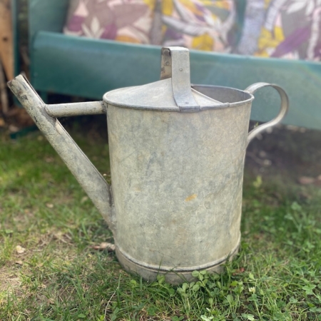 L'Arrosoir Galva 1960, un arrosoir en métal vintage patiné, est posé sur l'herbe avec un banc recouvert de coussins colorés en arrière-plan. Brocante 93