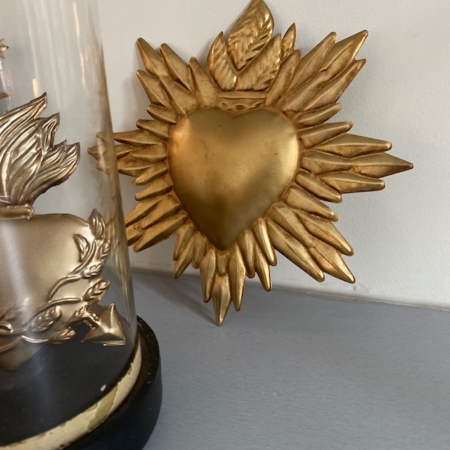 Le Coeur Jungle Or, présentant un symbole de cœur doré avec un design d'étoile rayonnante vintage, est placé à côté d'un dôme en verre décoratif sur une surface grise. Brocante 93