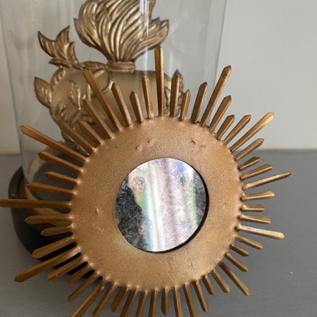 Le Miroir Mini Soleil Or, doté d'un centre circulaire réfléchissant avec des rayons semblables à ceux du soleil, est présenté devant une sculpture feuillue à l'intérieur d'un dôme de verre. Brocante 93