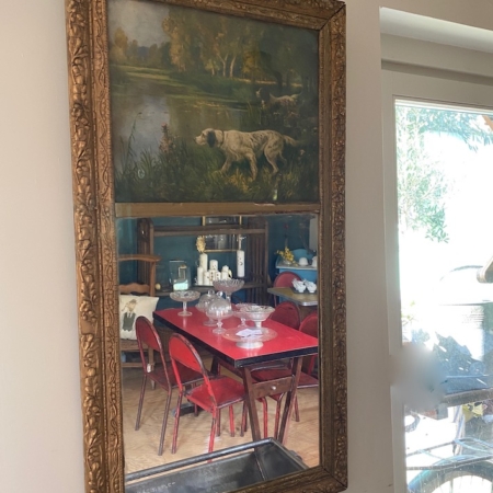 Le Miroir Trumeau 1940 VENDU, un grand miroir orné d'une peinture vintage représentant un chien au bord d'un étang au sommet, reflète un coin repas avec des chaises rouges et une table. Brocante 93