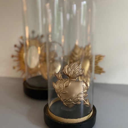 Deux sculptures Coeur Flèche Or, en or et en forme de cœur sacré, sont encastrées dans des dômes de verre, rayonnant d'un charme rétro avec des éléments décoratifs visibles en arrière-plan. Brocante 93