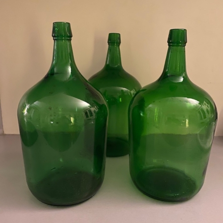 Trois bouteilles Dame Jeanne 1970 PM, caractérisées par leurs grands corps en verre vert et leurs cols étroits, sont posées sur une surface grise sur fond beige, dégageant un charme vintage. Brocante 93