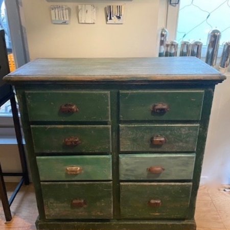 Le Meuble de Métier 1960, une commode rustique en bois vert à huit tiroirs, se dresse contre le mur et dégage un charme vintage, tandis que de petites pièces de décoration sont élégamment exposées sur des étagères au-dessus. Brocante 93