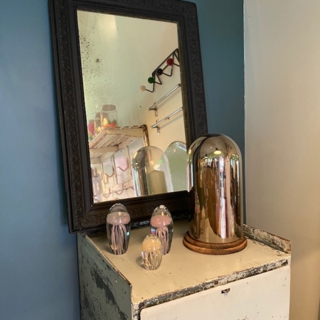 Un Miroir début XXème trône gracieusement aux côtés d'ornements décoratifs en verre sur une armoire vieillie contre un mur bleu sarcelle, évoquant un charme intemporel. Brocante 93