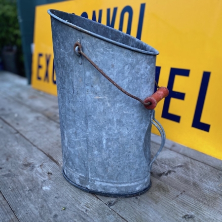 Le Seau à Charbon 1950 VENDU, avec sa surface en métal patiné et sa poignée en bois, est posé sur une table en bois, dégageant un charme vintage avec une enseigne jaune en arrière-plan. Brocante 93