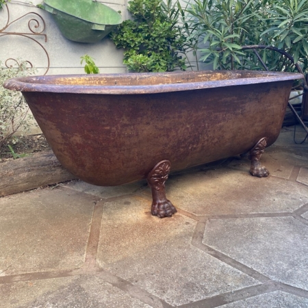 Une Baignoire Fonte 1900 VENDU, vieille et rouillée, trône à l'extérieur sur un espace pavé de pierres entouré de plantes luxuriantes. Brocante 93