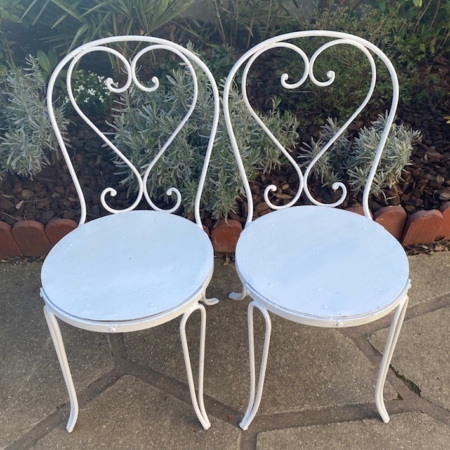 Une paire de chaises en fer forgé des années 1950, mettant en valeur l'artisanat du métal blanc avec des sièges circulaires et des dossiers au design complexe qui évoquent un charme vintage, orne le patio du jardin. Brocante 93