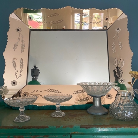 Un Miroir Bicolore 1950 VENDU aux motifs floraux gravés repose élégamment sur une commode verte, flanqué de bols et de vases en verre ornés. Brocante 93