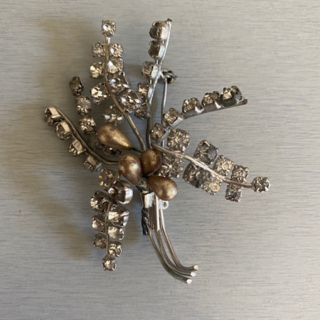 La Broche Rameaux Strass 1960 est une broche rétro en argent ornée d'accents de strass et de détails de perles, disposés dans un design de bouquet floral, parfait pour ajouter une touche de charme vintage. Brocante 93