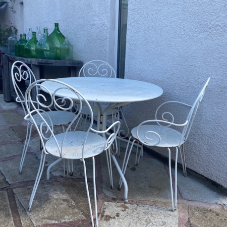 Une table ronde blanche vintage et ses quatre chaises assorties, faisant partie de la collection « Table & Chaises de Jardin VENDU », sont assises sur une terrasse en béton près d'un mur orné de grandes bouteilles en verre vert sur un rebord, dégageant un charme rétro intemporel. Brocante 93