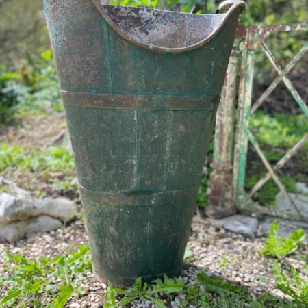 Une Hotte à Vendange vintage 1960 VENDU se dresse debout sur l'allée de gravier, entourée d'un feuillage vert luxuriant. Brocante 93