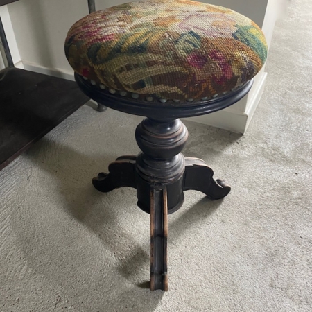 Le Tabouret de Piano début XXème siècle, avec son assise tapissée de motifs floraux, repose élégamment sur un sol recouvert de moquette près d'une étagère en métal. Brocante 93