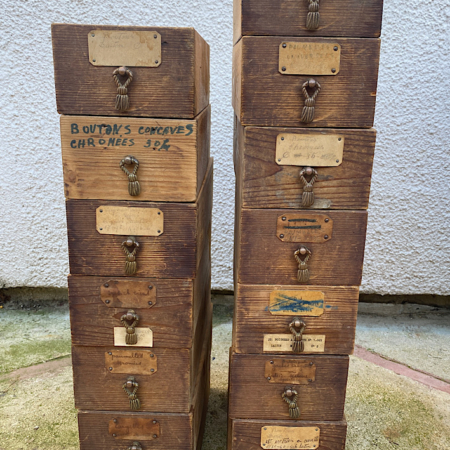 Deux piles de tiroirs vintage en bois avec poignées en métal, étiquetés avec des textes variés, posés sur une surface en béton évoquent une charmante esthétique rétro. Découvrez ces trouvailles uniques chez Brocante 93 sous le nom de "Lot de Tiroirs 1960 MM VENDU". Brocante 93