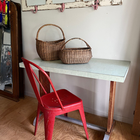 Une chaise rouge rétro trône à côté de la Table de Bistrot 1960, ornée de deux paniers tressés, dans une pièce rustique vintage dotée de crochets muraux et d'un plancher en bois. Brocante 93