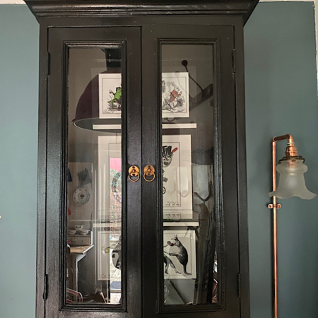 La Vitrine Murale 1940 VENDU, une armoire en bois foncé avec des portes vitrées, présente des tableaux encadrés à l'intérieur, tandis qu'une lampe vintage se trouve à droite. Brocante 93