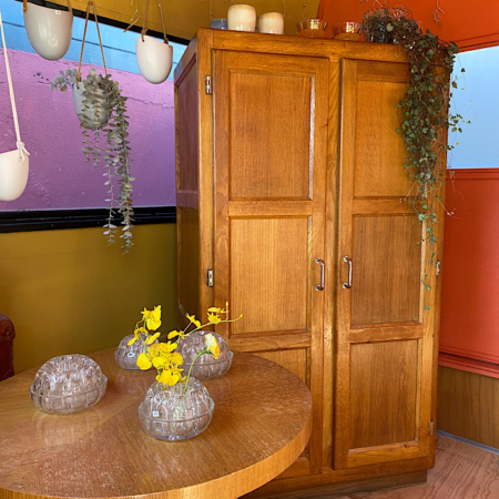 Chambre cosy au mobilier chaleureux en bois mettant en scène l'élégante Armoire Parisienne 1960 VENDU, une table ronde ornée d'un décor en verre vintage, et des plantes suspendues et sur un meuble. Brocante 93