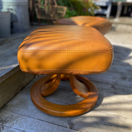 Une Paire de Poufs Cuir 1980 VENDU, avec son revêtement en cuir couleur caramel et sa base en bois, repose gracieusement sur une terrasse en bois, projetant des ombres qui rehaussent son charmante ambiance vintage. Brocante 93