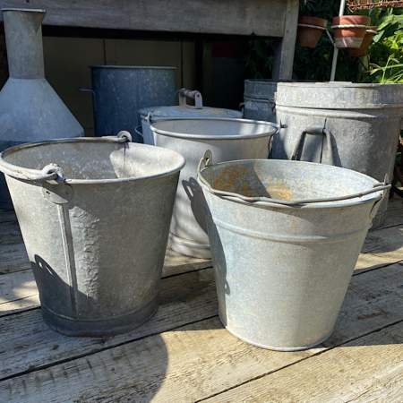 Quatre seaux Seaux en galva 1960 sont posés sur une surface en bois à l'extérieur, évoquant un charme rétro distinct, avec des récipients en métal vintage supplémentaires en arrière-plan. Brocante 93